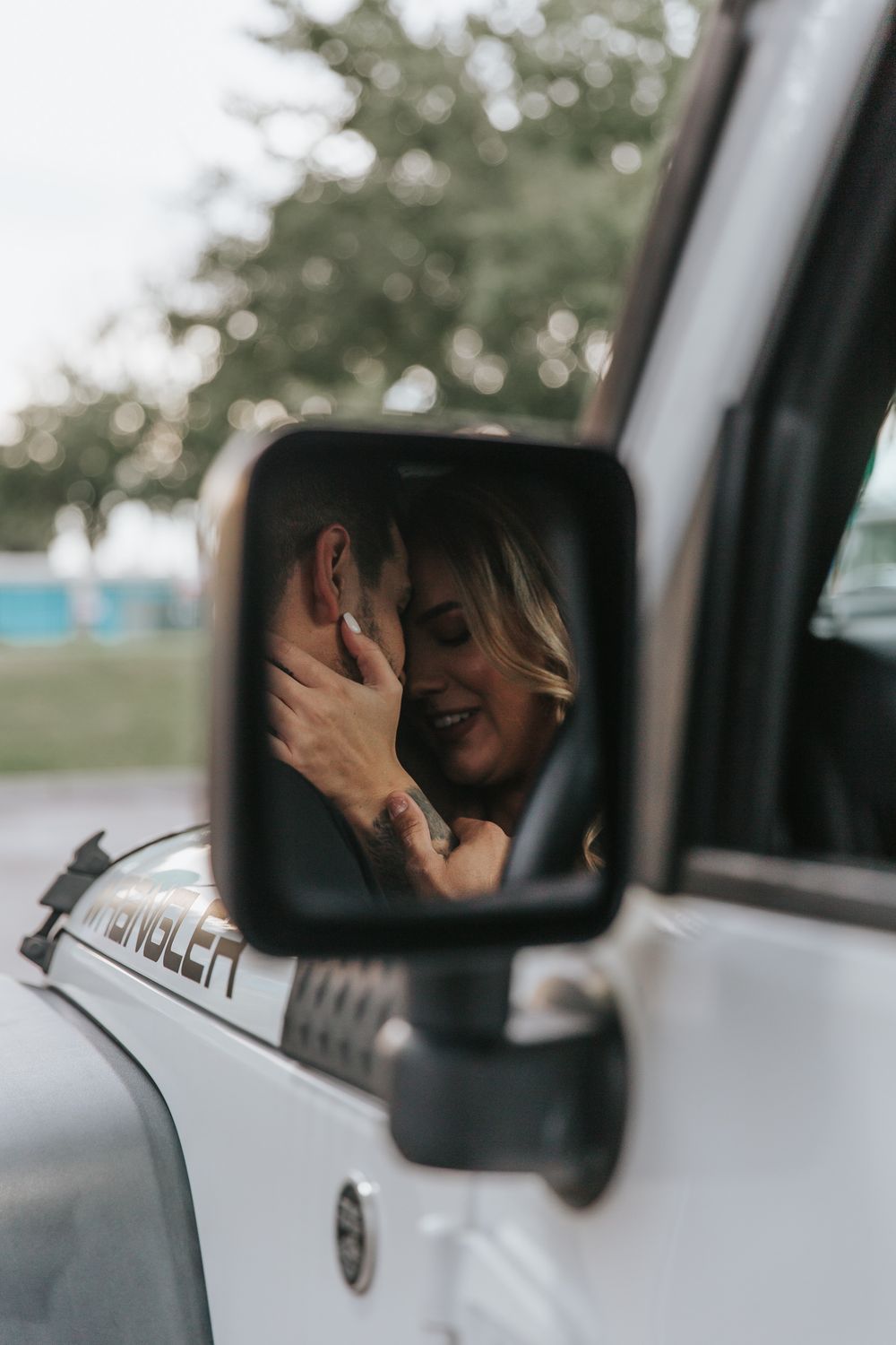 A romantic couple shares an intimate moment reflected in a car's side mirror.