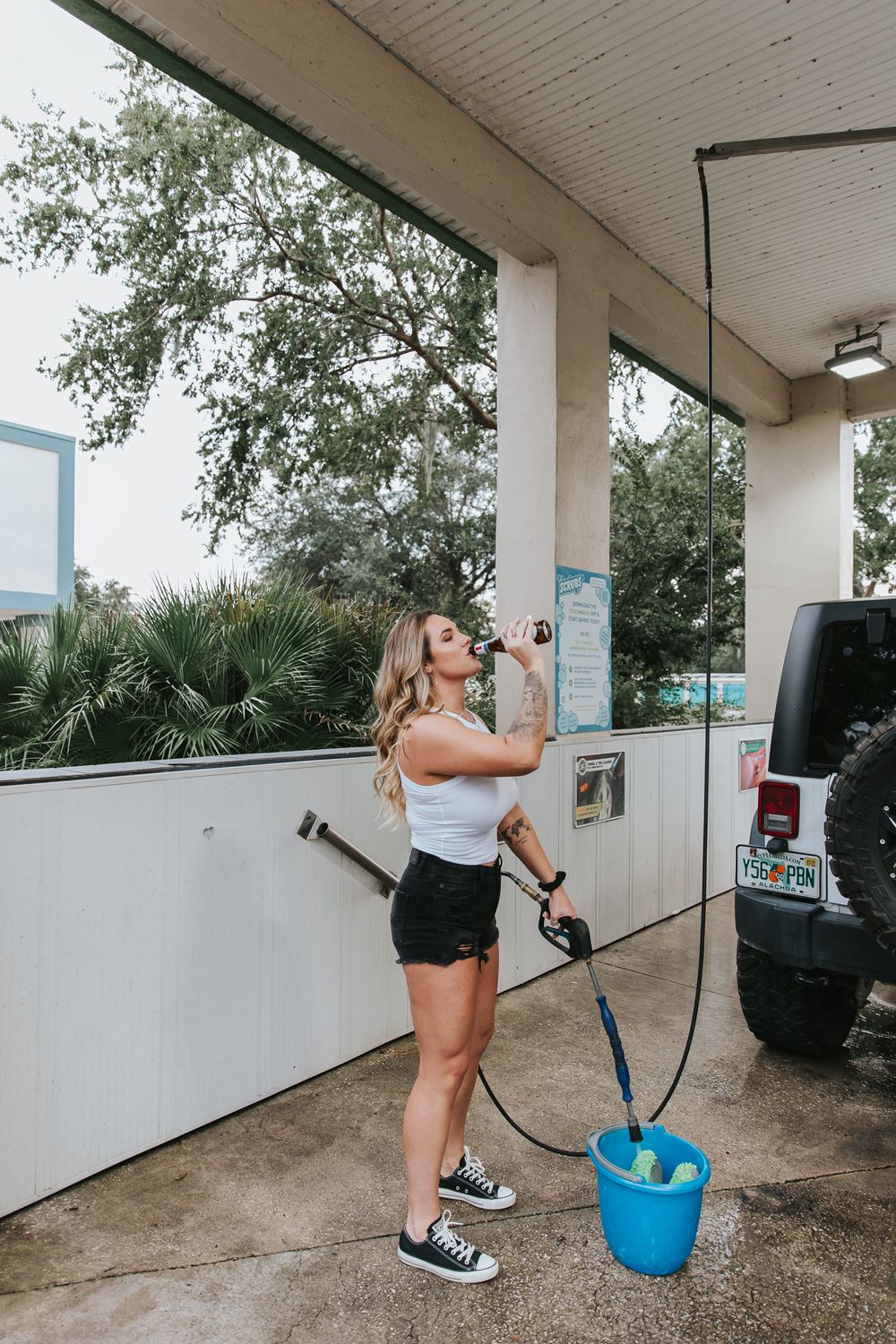 A couple playfully washing a black Jeep Wrangler under a covered area on a sunny day.