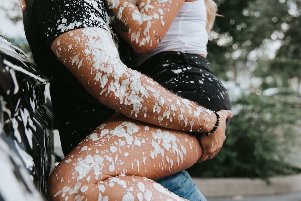 A person wearing jeans sits on a rustic wooden bench covered in white paint flakes in an outdoor setting.