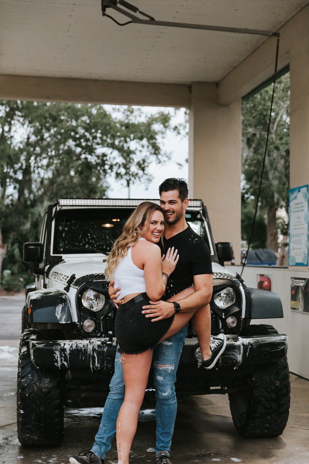 A couple poses playfully at a car wash with a black off-road vehicle in the background.