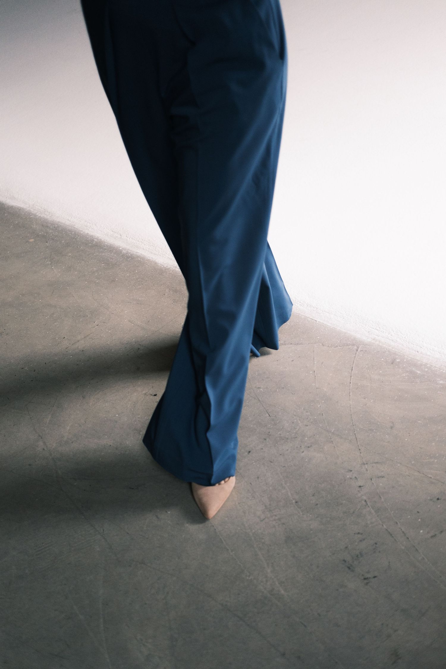 Close-up view of navy blue pants and bare feet walking on a concrete floor in a minimalist setting.