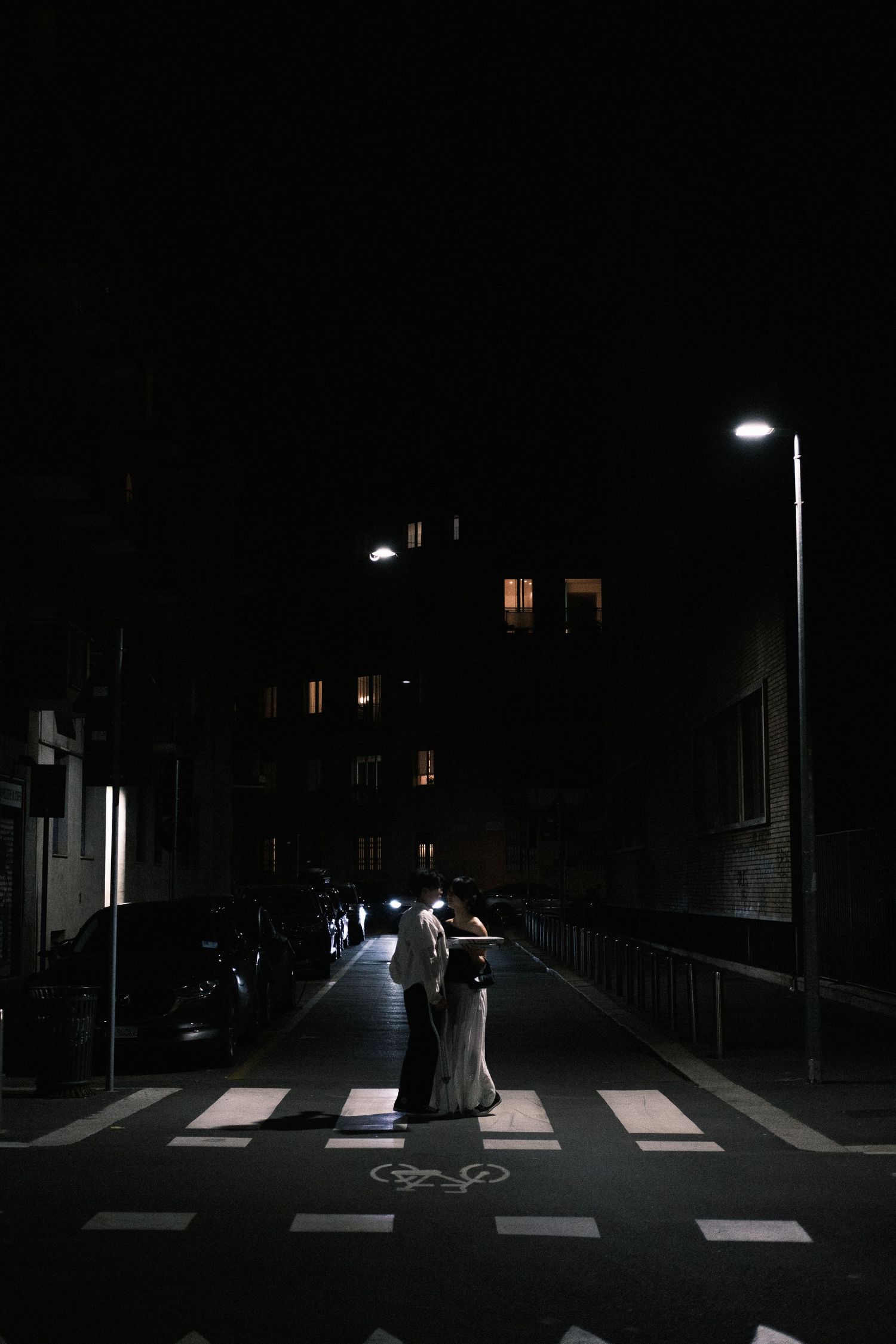 A figure in a flowing white dress crosses a dimly lit crosswalk at night under a street lamp's glow.