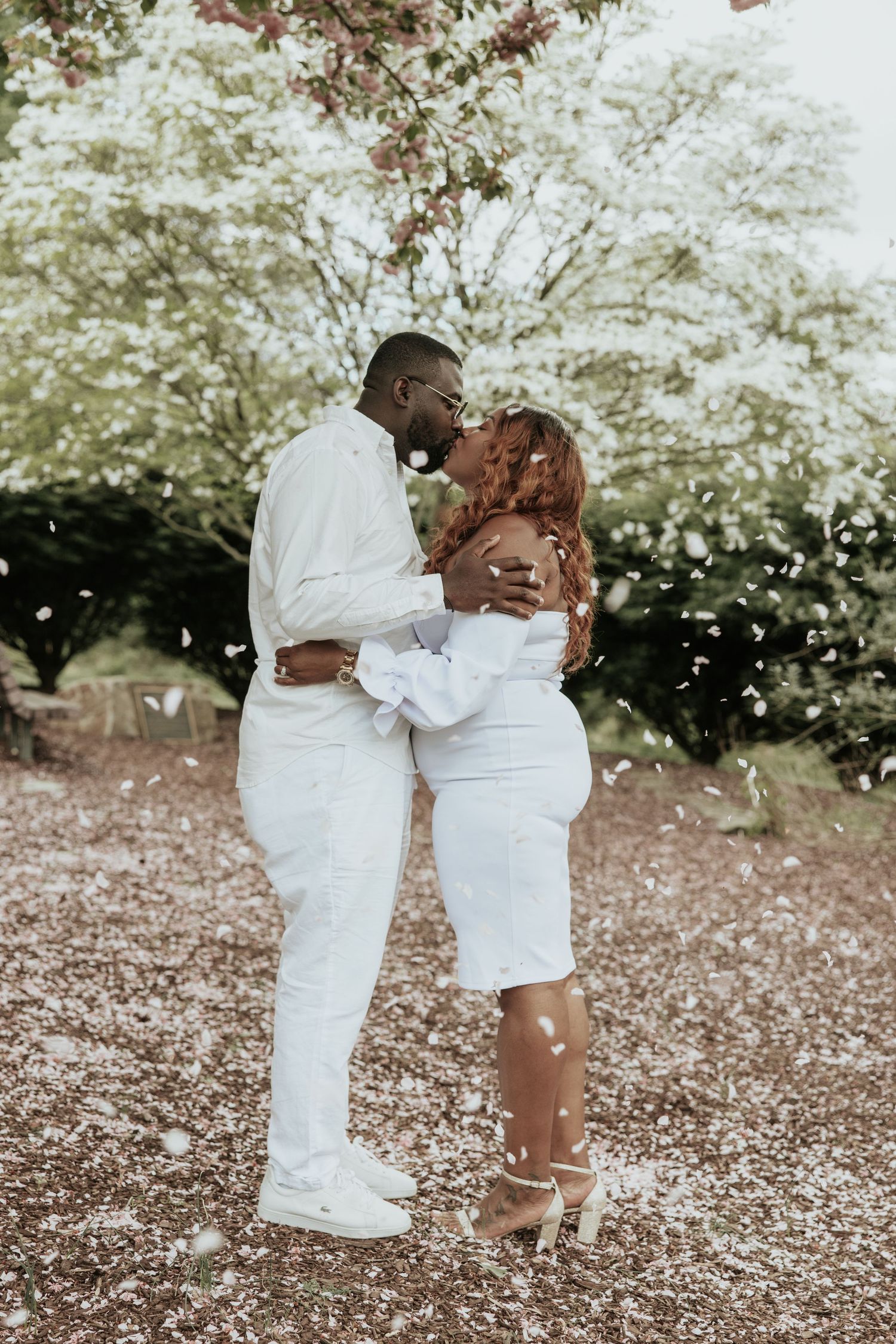 A romantic couple in matching white outfits embrace under blooming trees with petals falling around them.