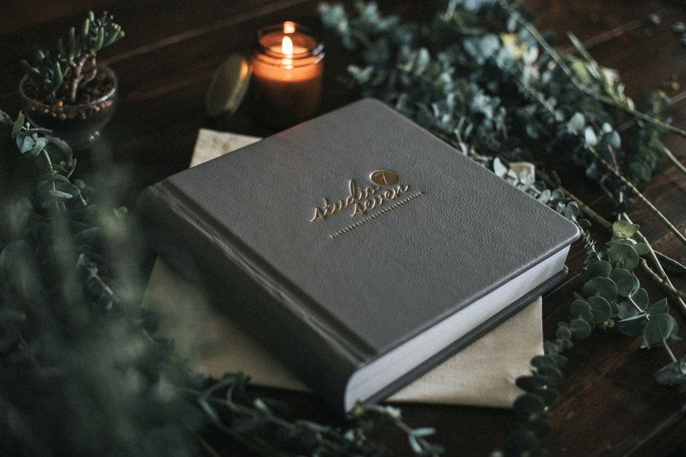 Moody still life of dark leather-bound books and candles with greenery on wooden surface.