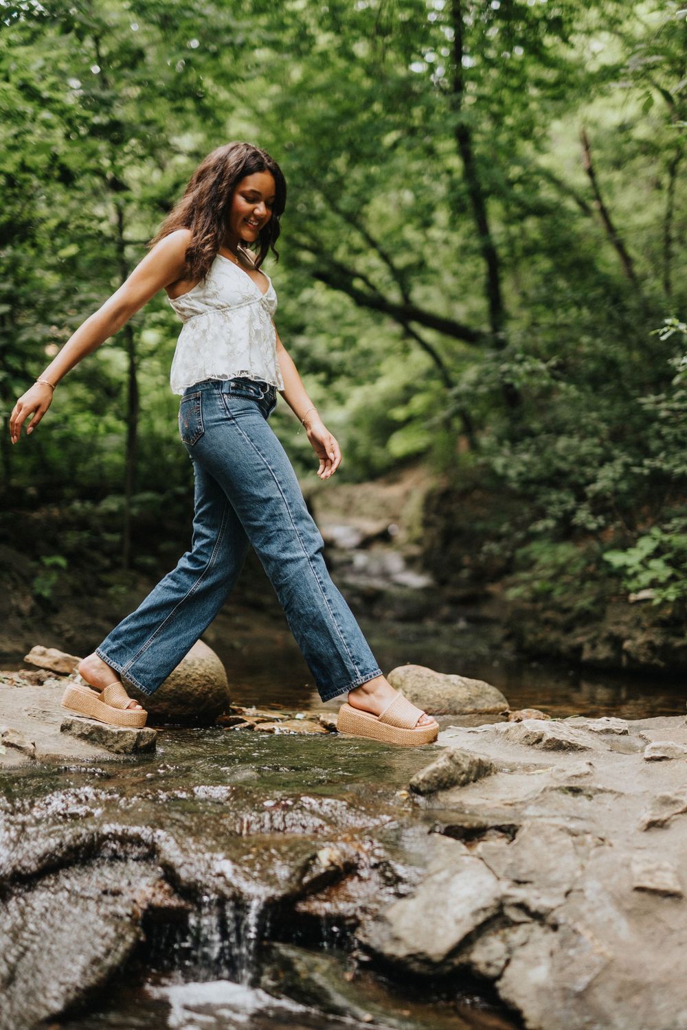 A person in denim jeans carefully steps across wet rocks in a forest stream.