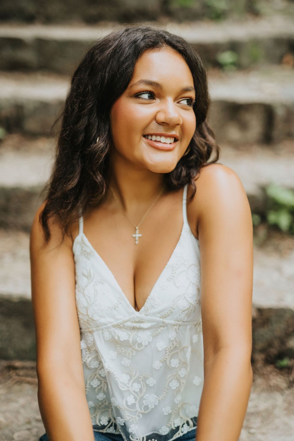 A bride in a white lace dress with floral appliques smiles while sitting outdoors on stone steps.