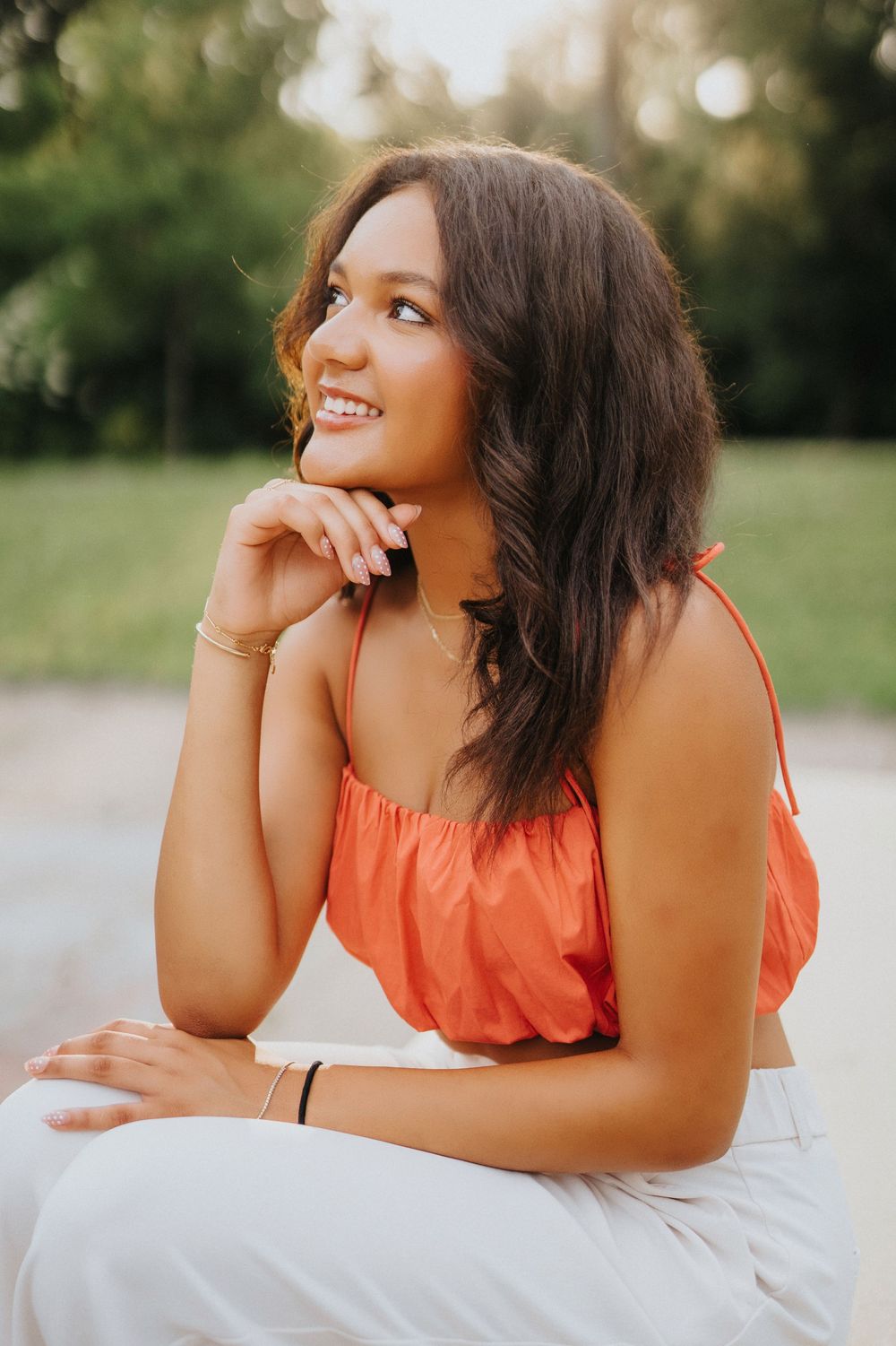 A person in an orange tank top and white pants smiles while posing outdoors during golden hour.