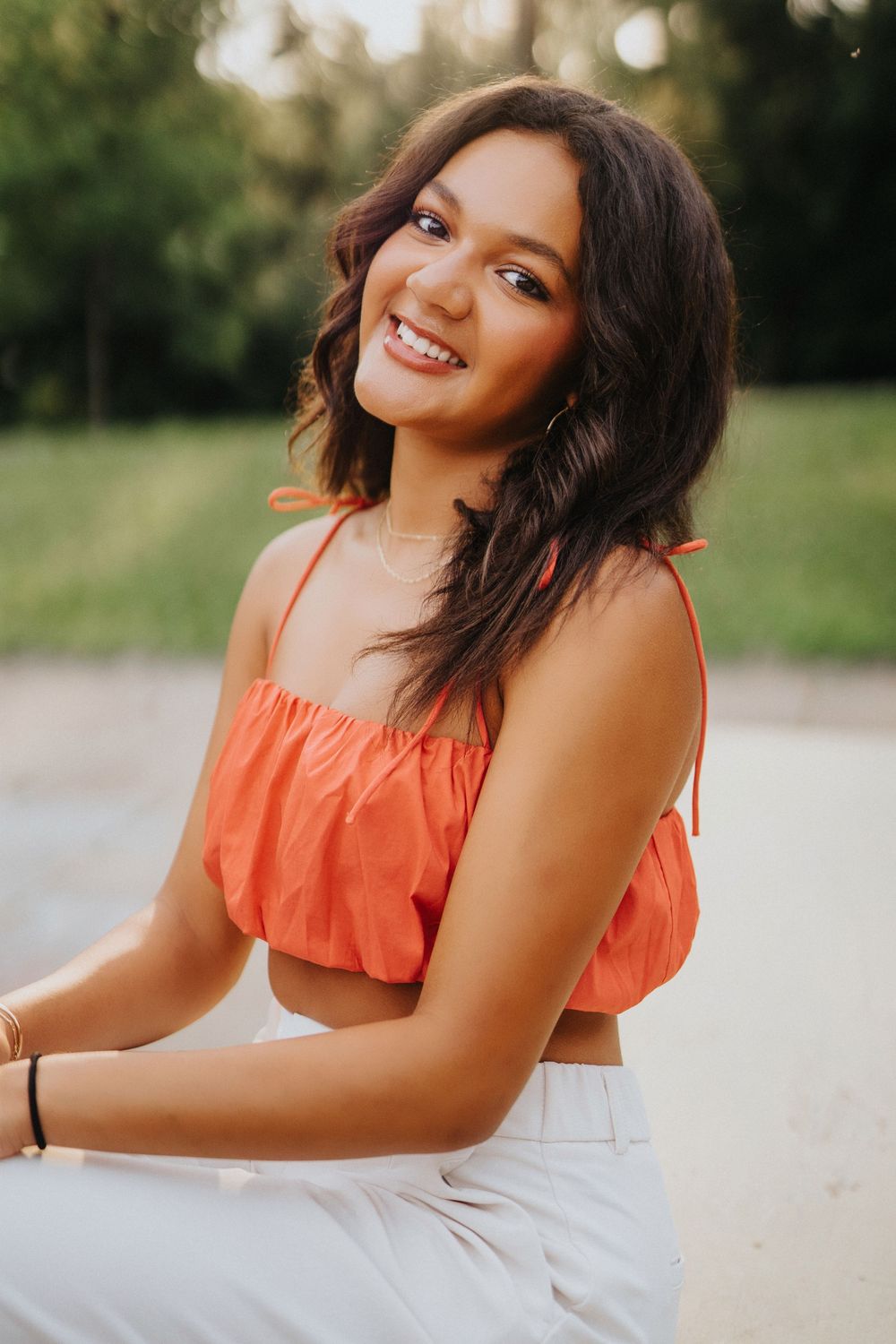 A woman in a coral crop top and white pants smiles warmly while posing outdoors during golden hour.