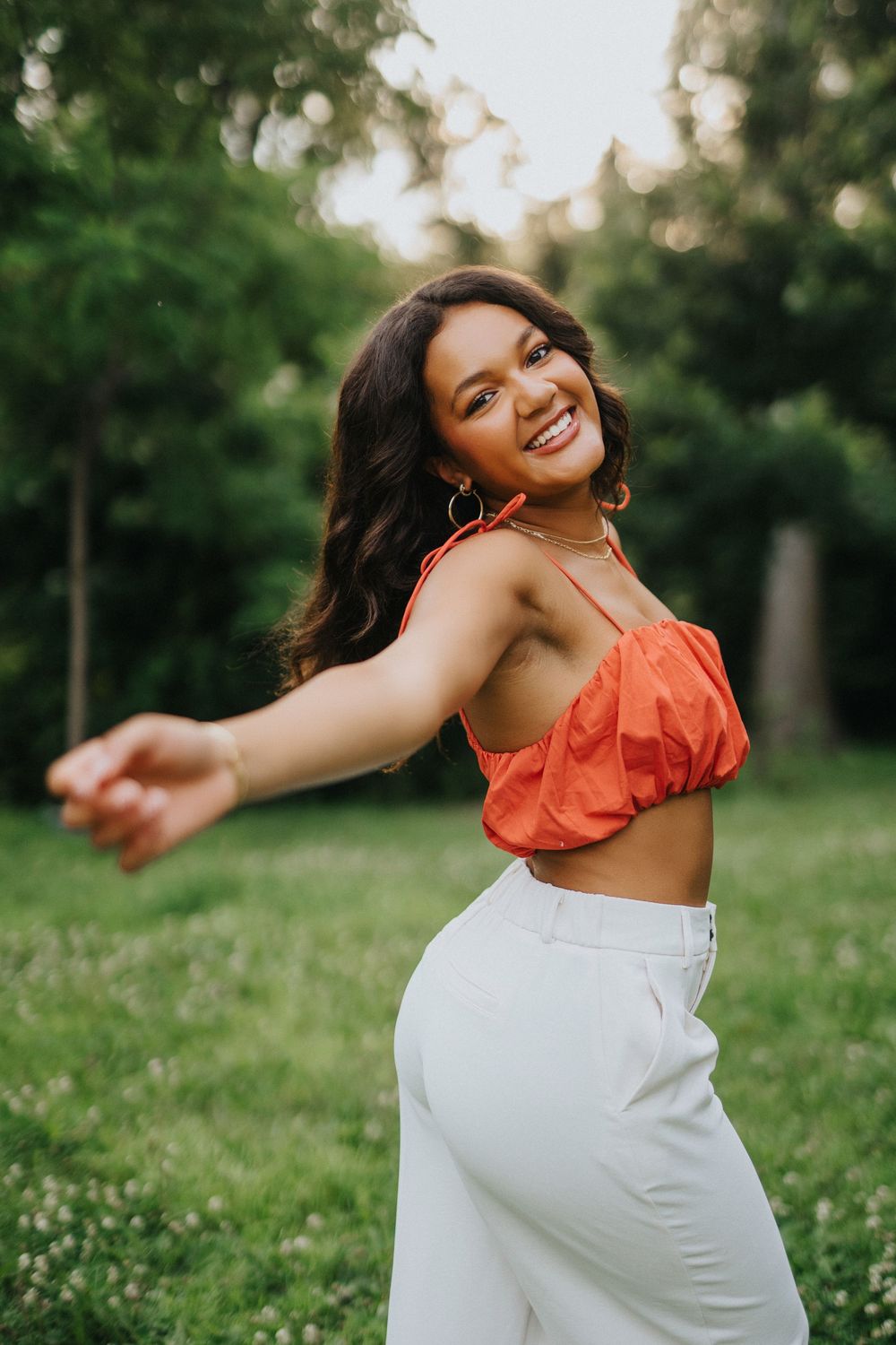 A joyful dance sequence in white pants and orange top captured against a lush green outdoor backdrop.