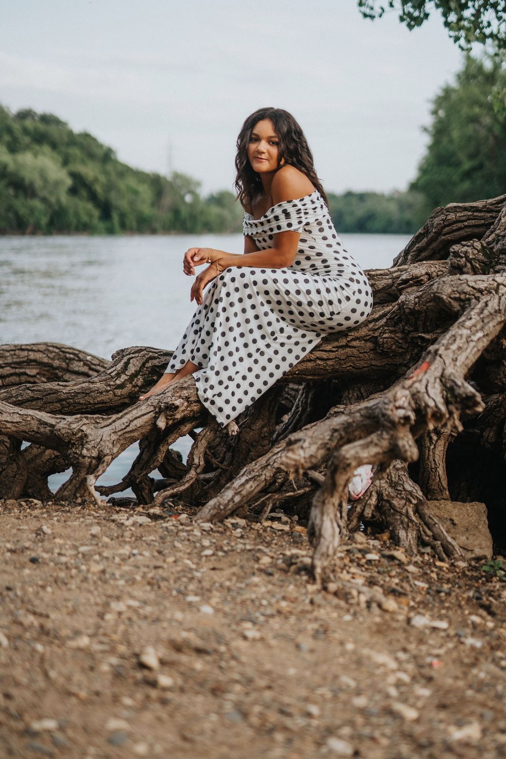 A person in a polka dot dress poses gracefully on driftwood by a riverbank during golden hour.