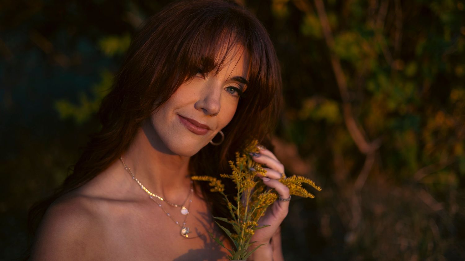 A woman in a dark dress poses among autumn foliage and flowers at sunset creating a dreamy outdoor portrait.