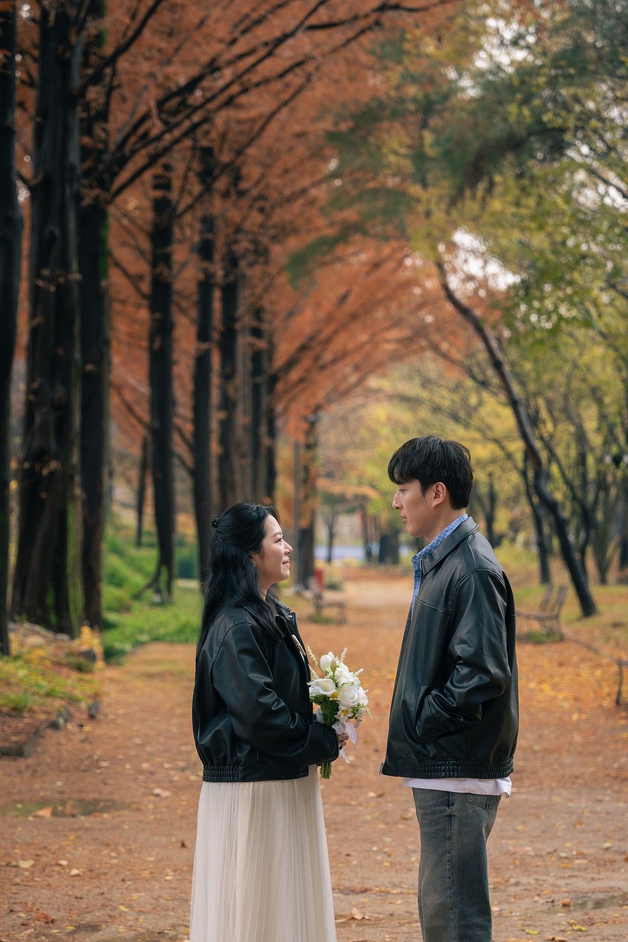A couple in formal attire walks along a tree-lined autumn path covered in fallen orange leaves.