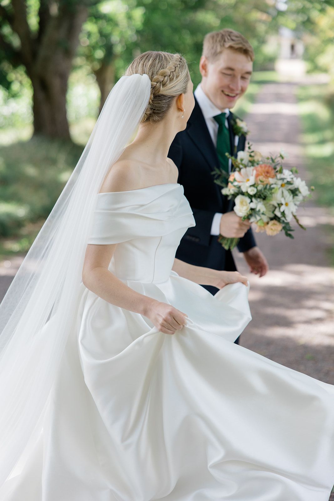 A bride in an elegant white off-shoulder gown and veil walks along a tree-lined path with a bouquet of flowers.