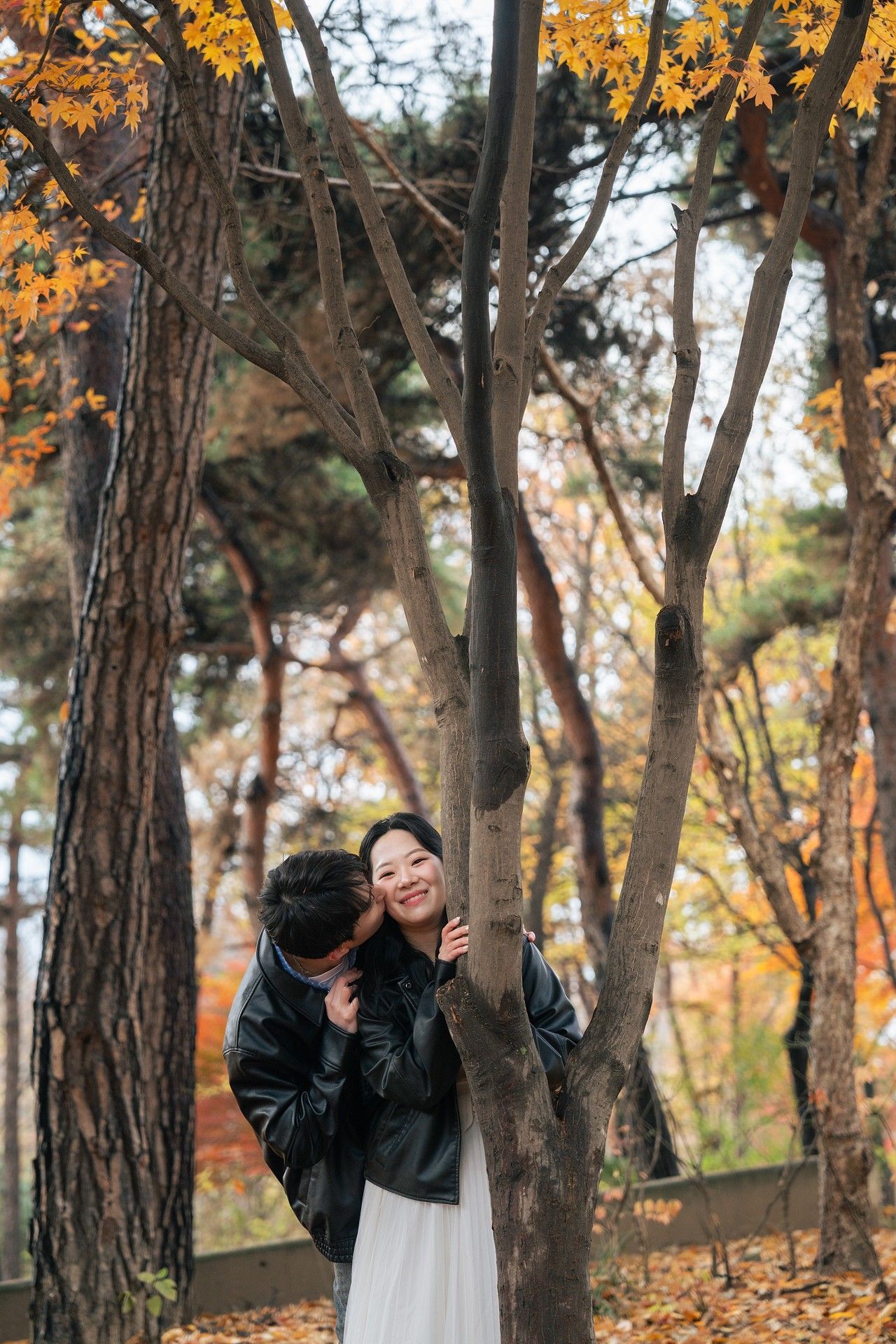 Person in black jacket poses playfully among autumn trees with golden leaves in a forest setting.