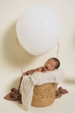 Newborn baby posed in woven basket with white balloon accents against cream background.