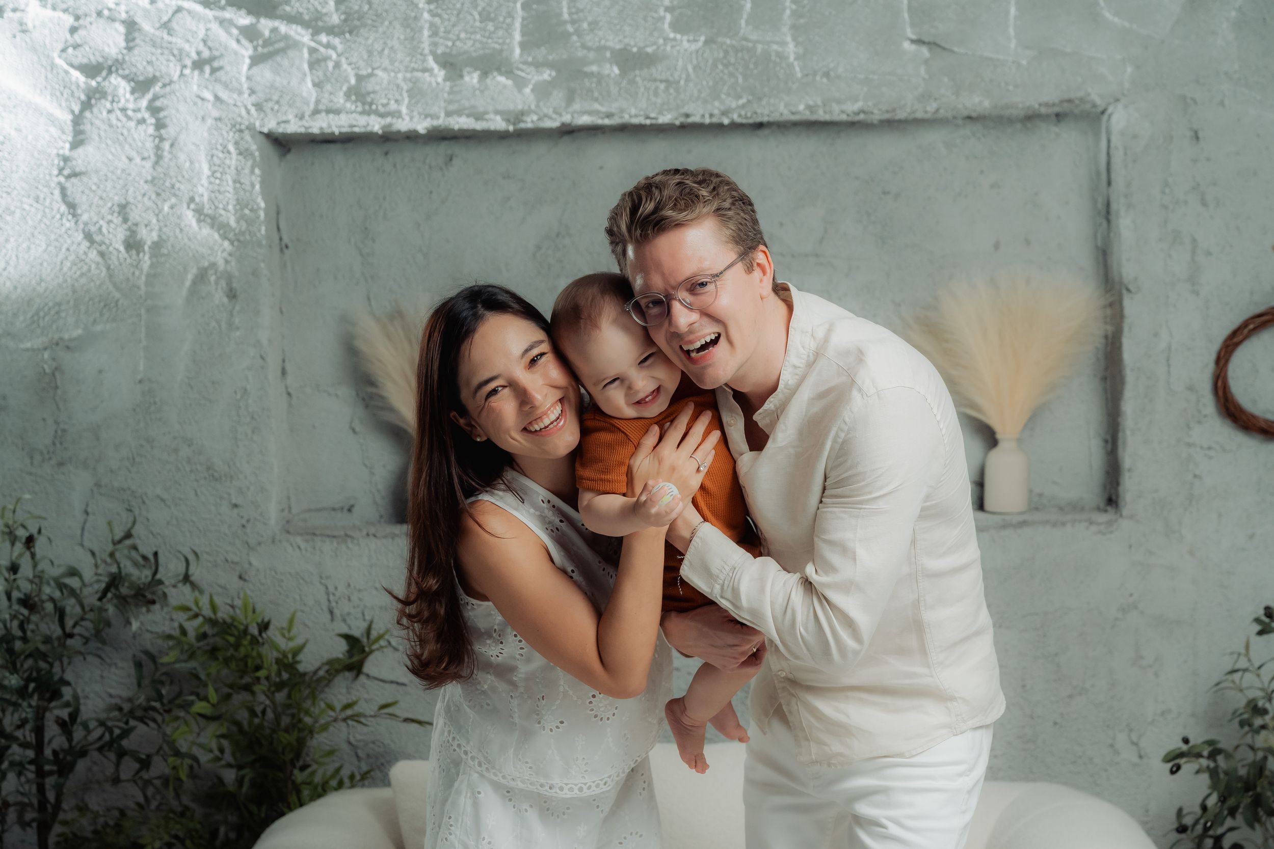 Family portrait of three people wearing white outfits embracing and laughing together against a grey wall.