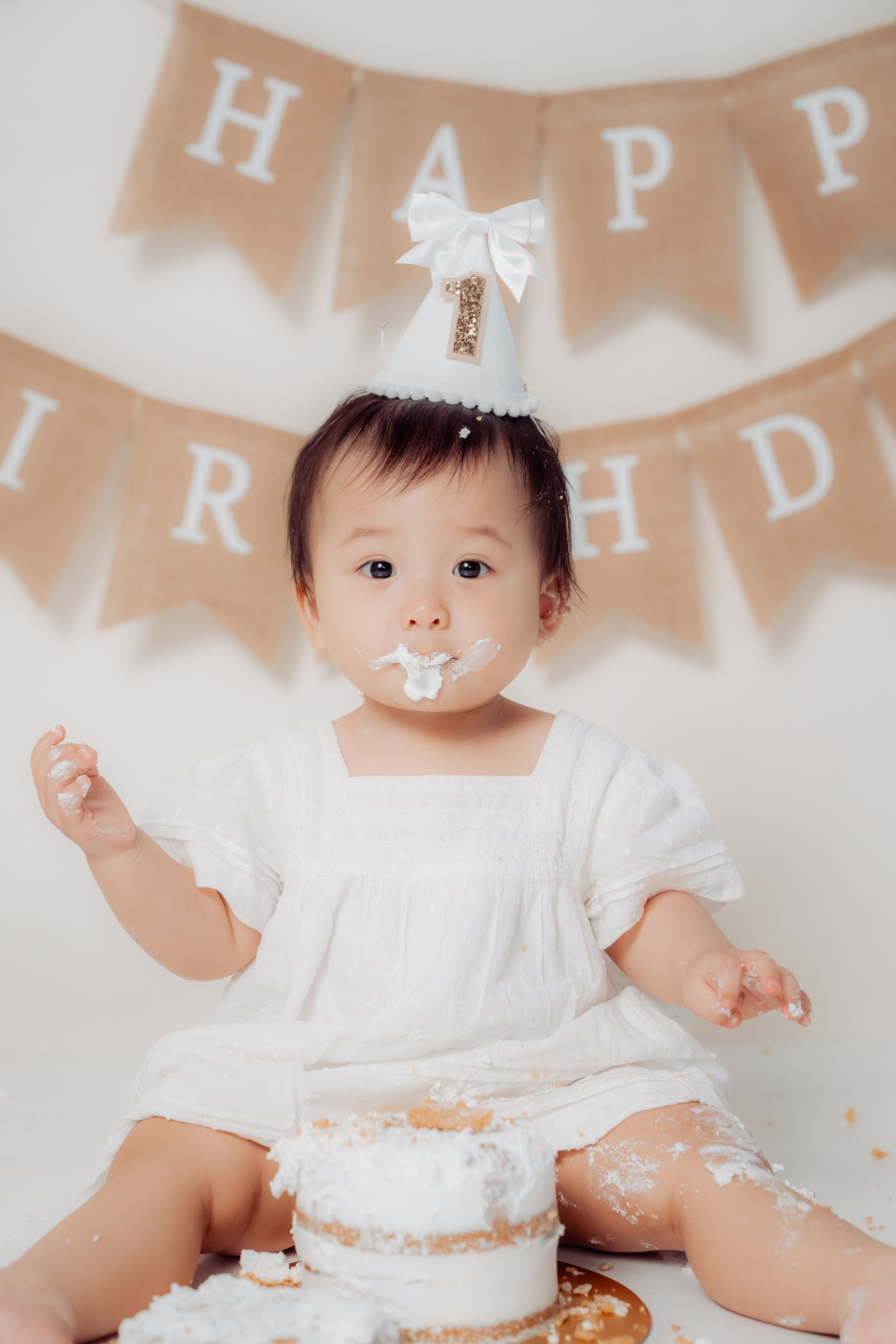 Baby in white dress having a cake smash photo session with 'HAPPY' banner decoration in the background.