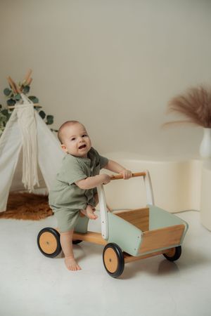 Toddler in sage green outfit playing with a wooden push cart toy near a small teepee tent.