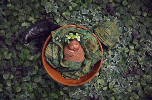 Newborn photography featuring baby nestled in terracotta pot surrounded by lush green foliage.