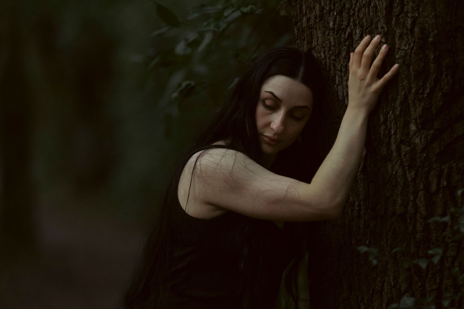 A figure in dark clothing leans against a tree trunk in a moody forest setting with dark atmospheric lighting.