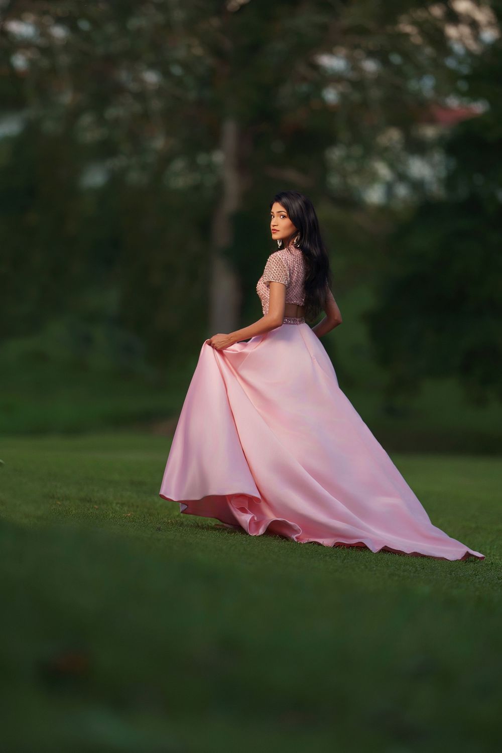 A flowing pink ball gown dress creates an elegant silhouette against a lush green outdoor backdrop.