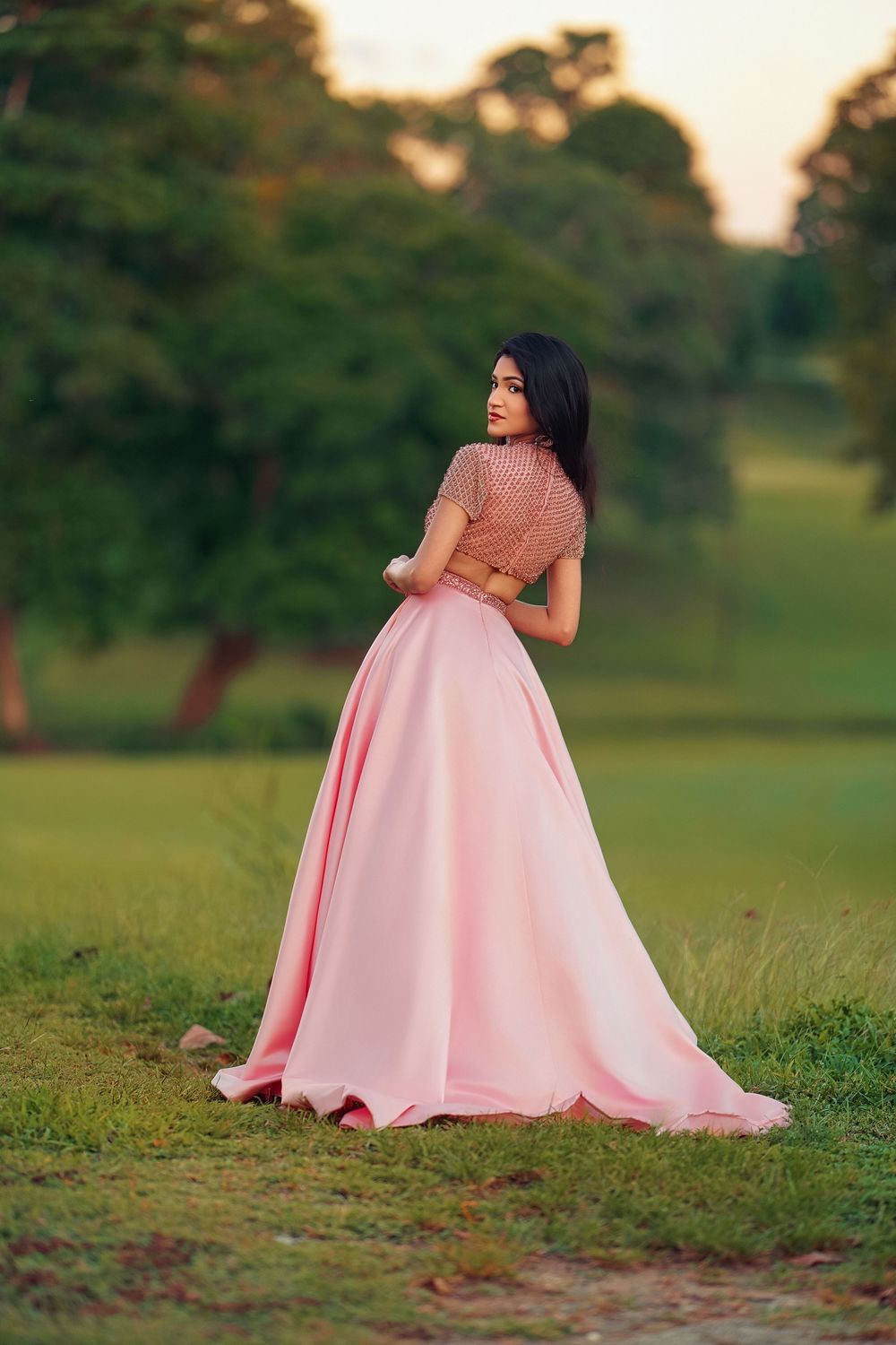 A pink ballgown dress flows elegantly across a grassy field during golden hour sunset.