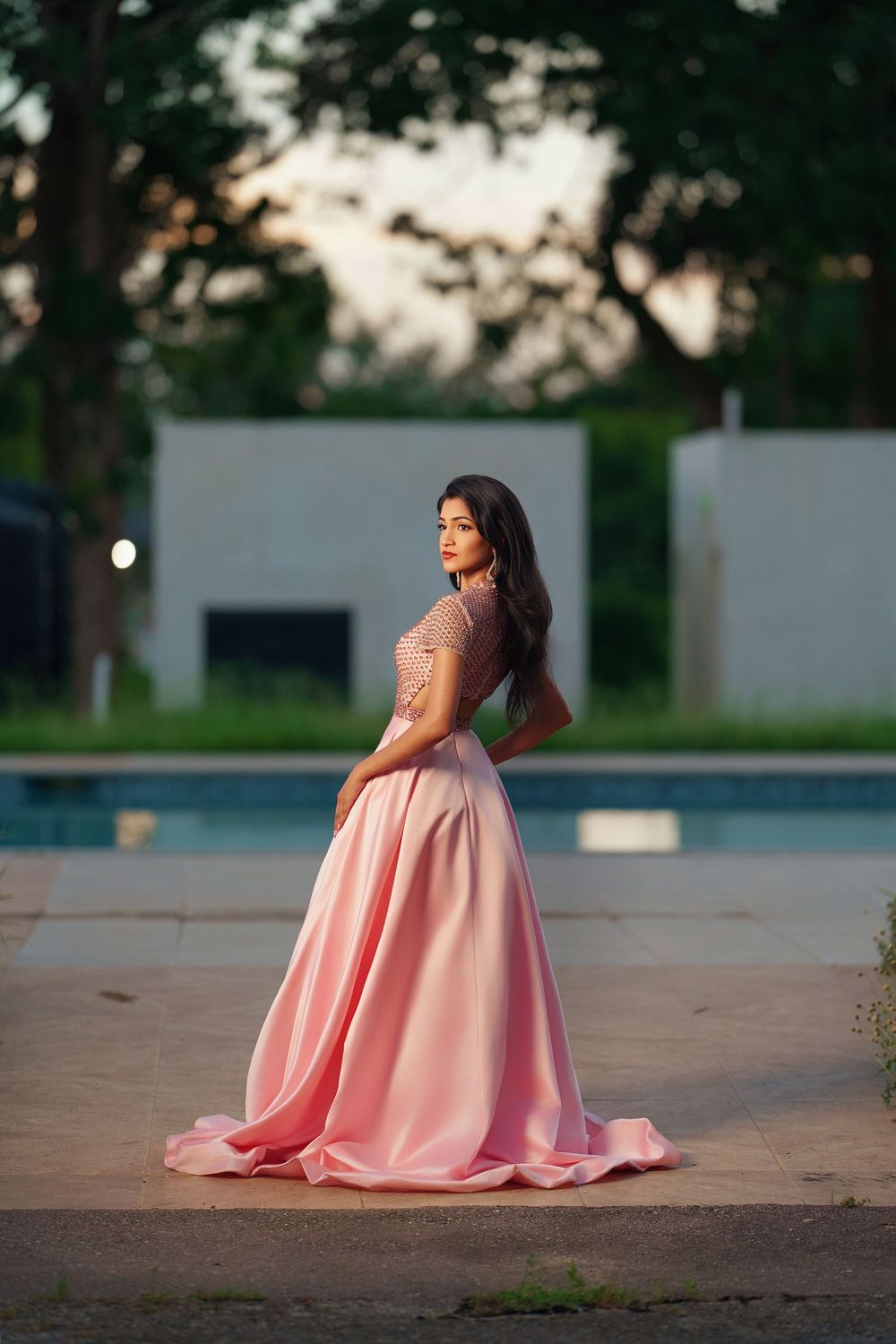 A flowing pink evening gown is captured in an outdoor photoshoot at dusk with trees in the background.