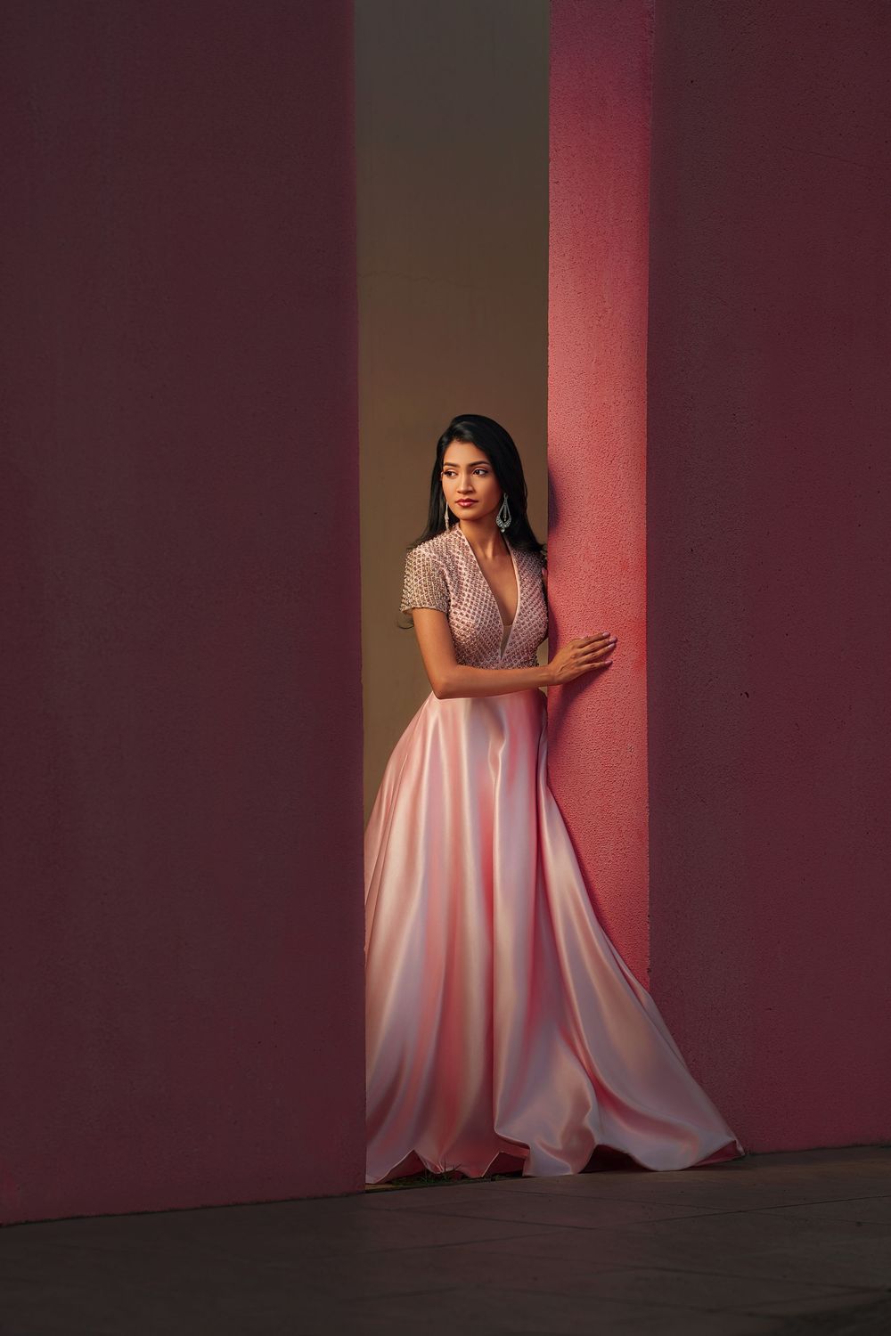 A pink evening gown creates dramatic shadows and movement against a dark burgundy wall in indoor lighting.