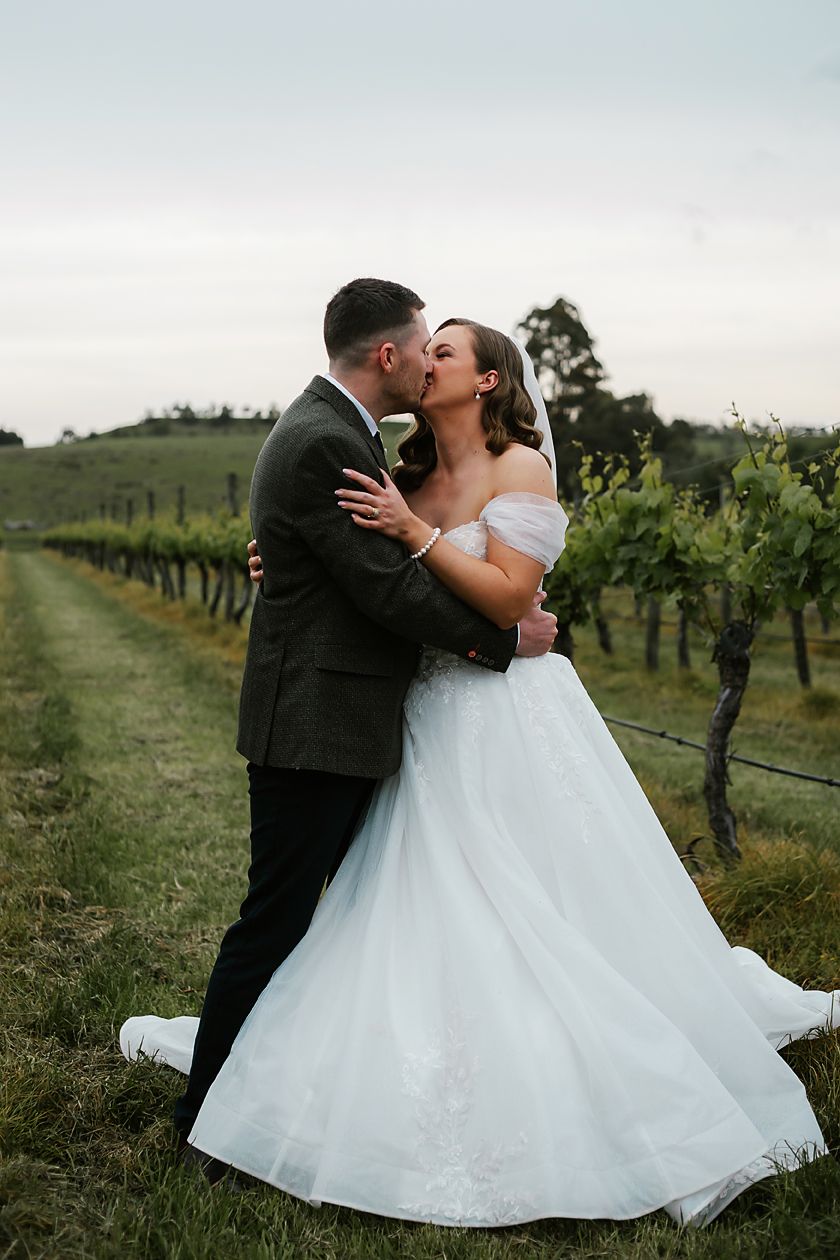 A romantic wedding couple shares intimate kisses among the vineyard rows at sunset.