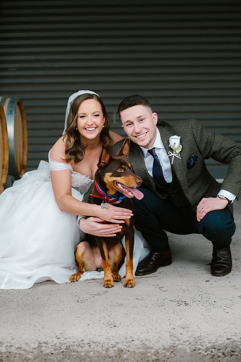 A couple in wedding attire poses with their brown dog wearing a bowtie against a rustic wall background.