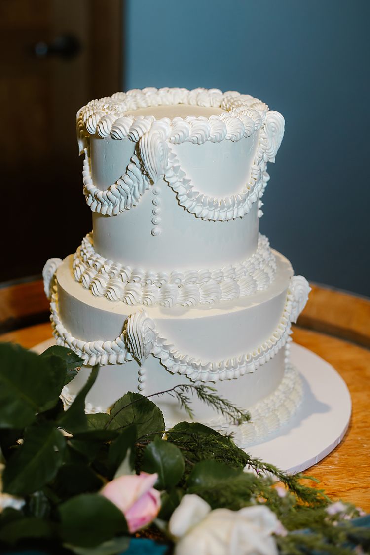 A white wedding cake with delicate piped frosting details and ruffled edges displayed on a wooden table with floral decor.