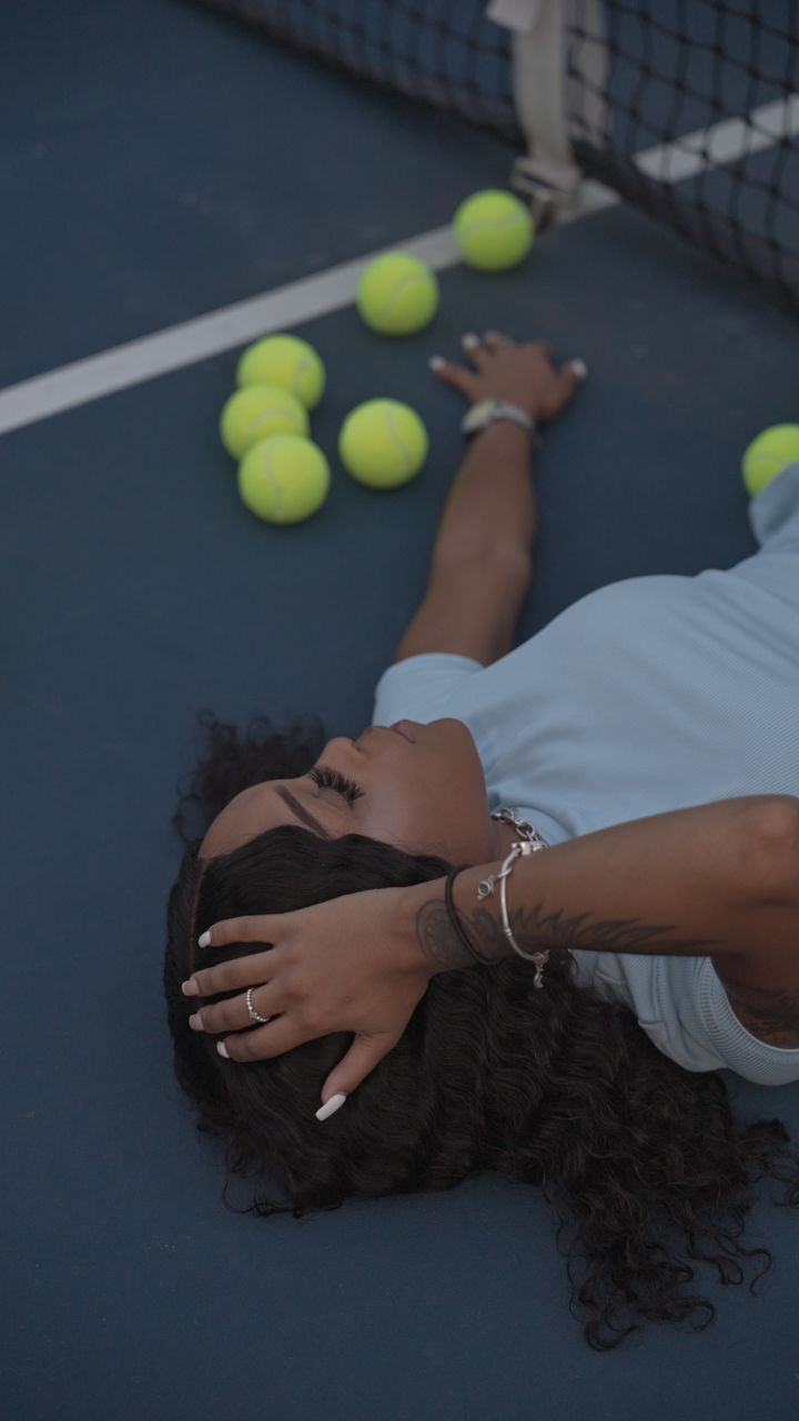 Person lying on tennis court with yellow tennis balls scattered nearby.