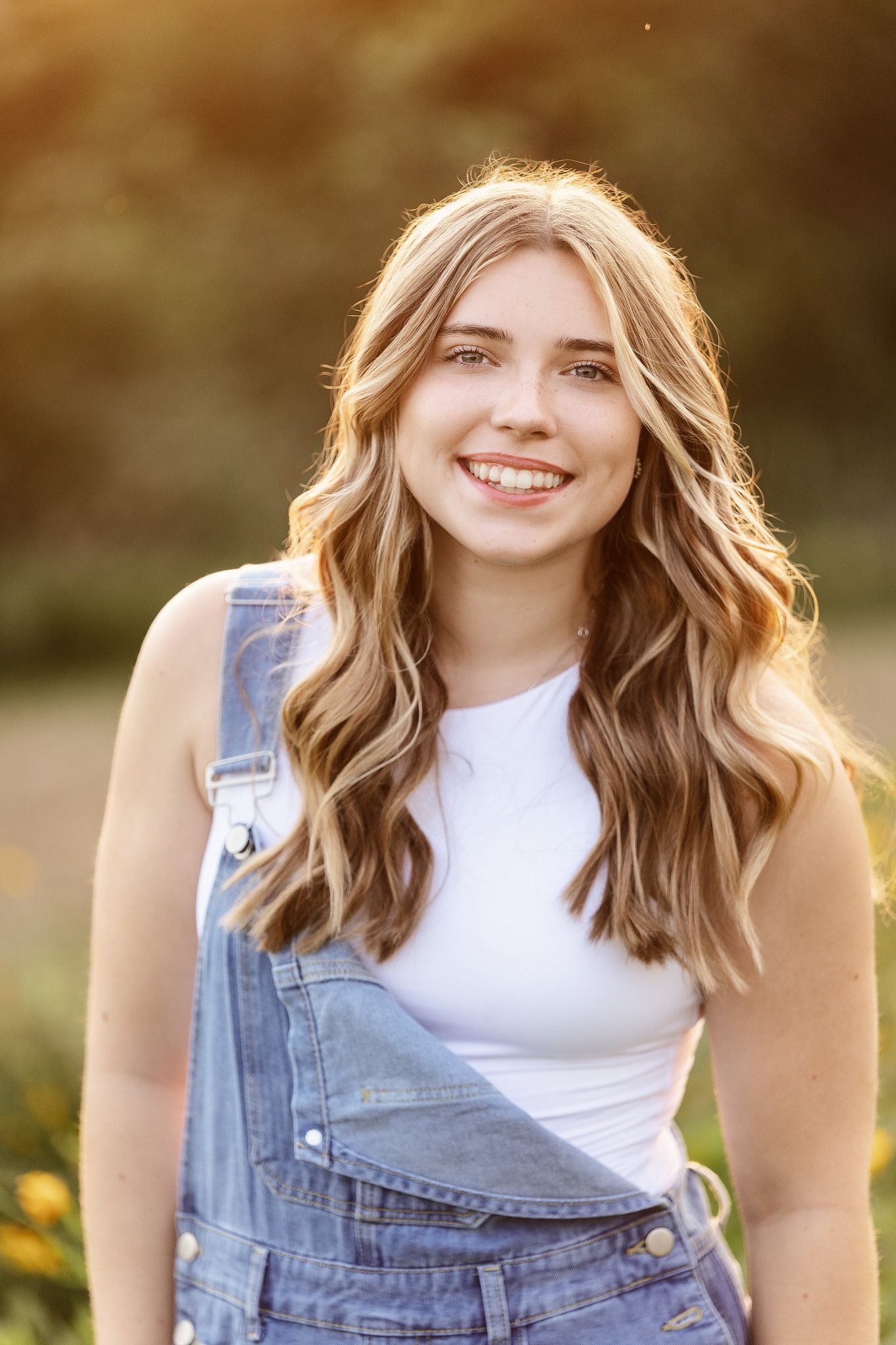 A person in denim overalls and white top poses outdoors during golden hour with natural sunlight.