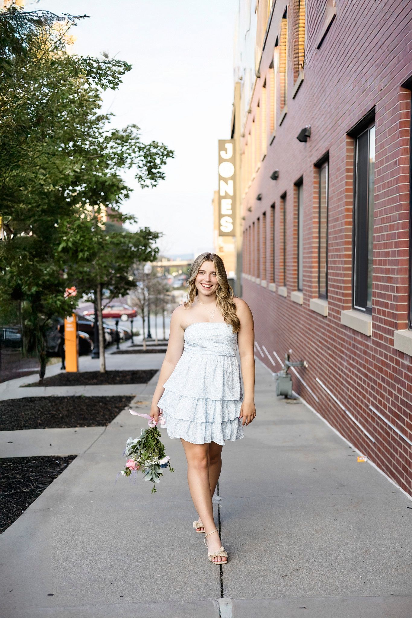 A person in a white ruffled dress poses against a brick wall in an urban setting, holding a bouquet of flowers.