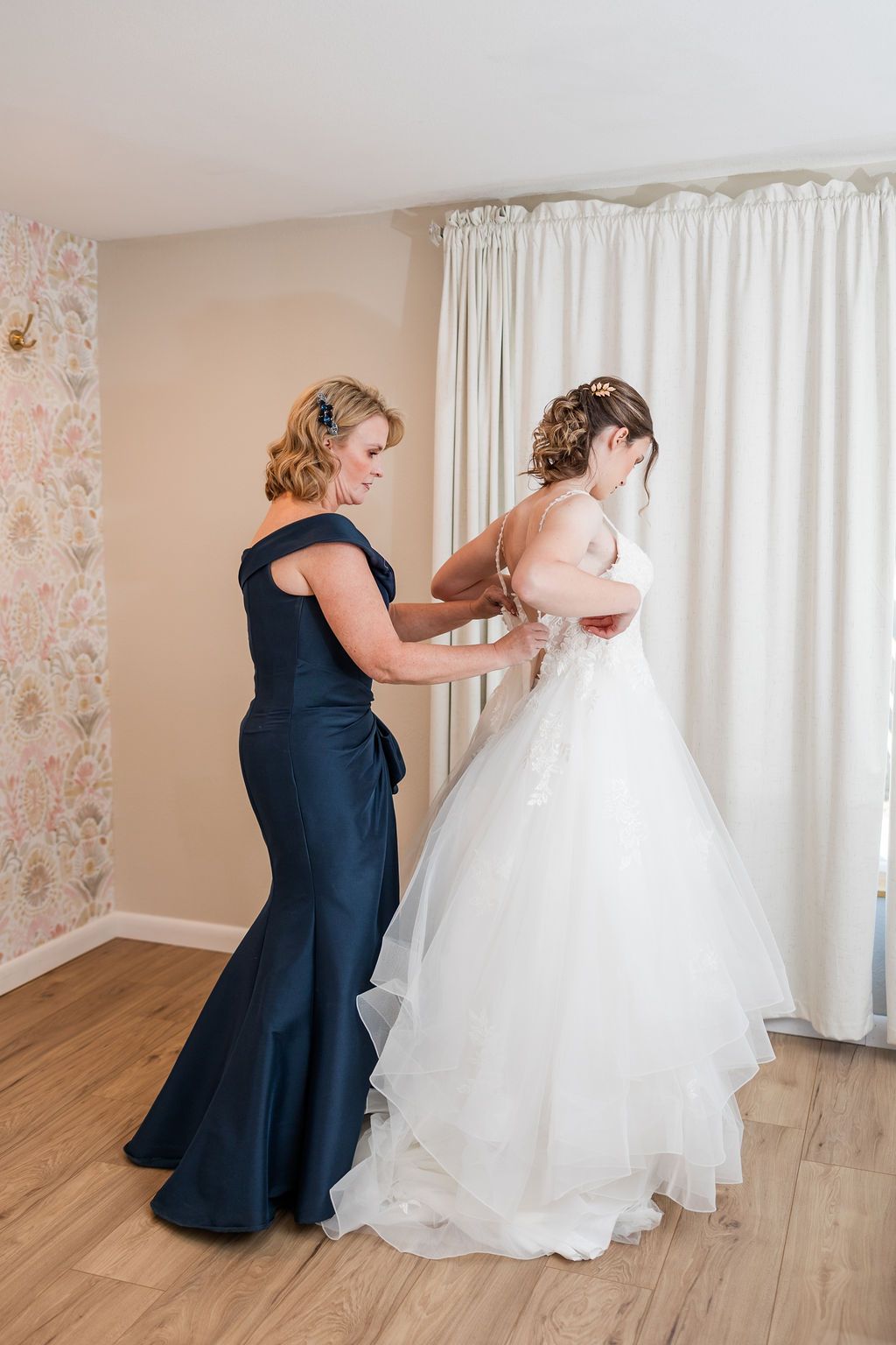 A bridesmaid in a navy dress helps adjust the bride's white ballgown wedding dress in a bright room.