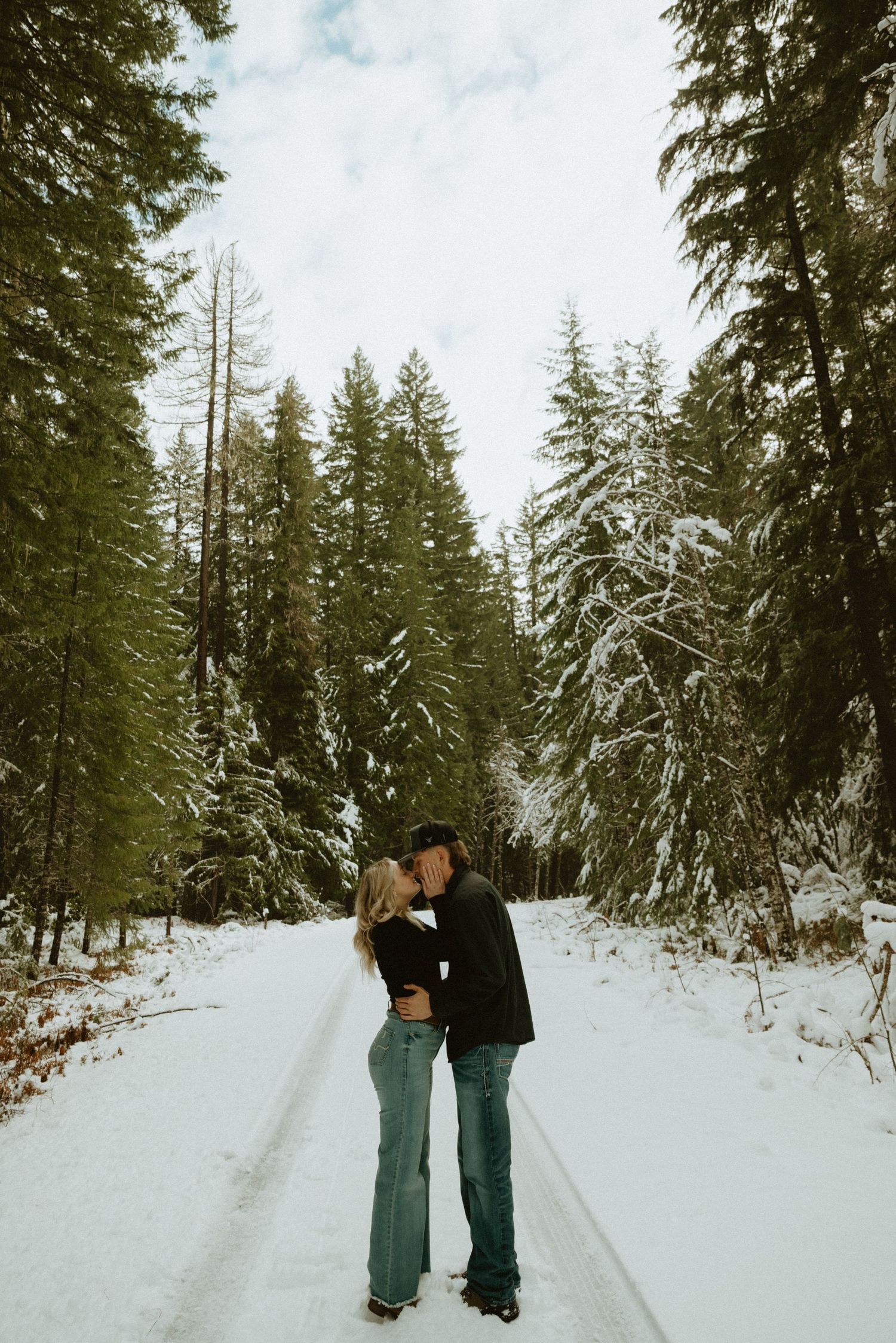 A couple playfully dances together in a snowy forest setting wearing casual winter attire.