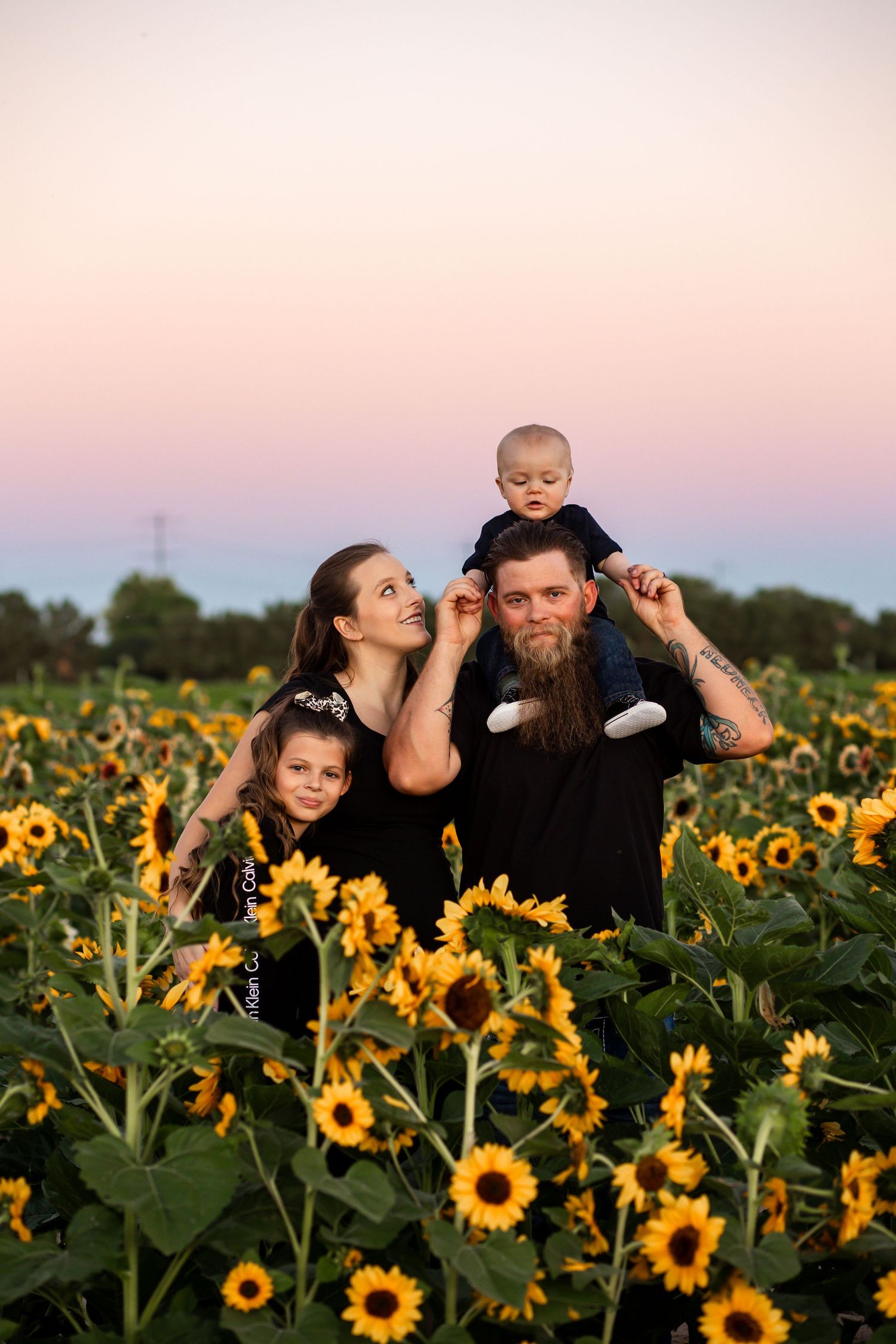 Family of four in black outfits stands together in a sunflower field at dusk.