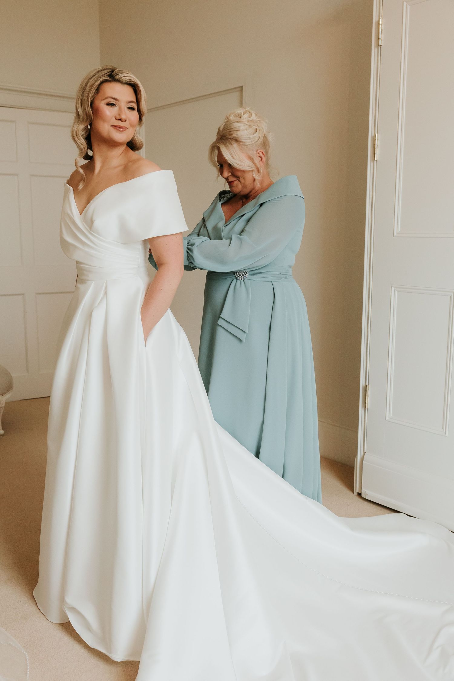 A bride in an elegant white wedding dress gets help with final adjustments in a softly lit room.