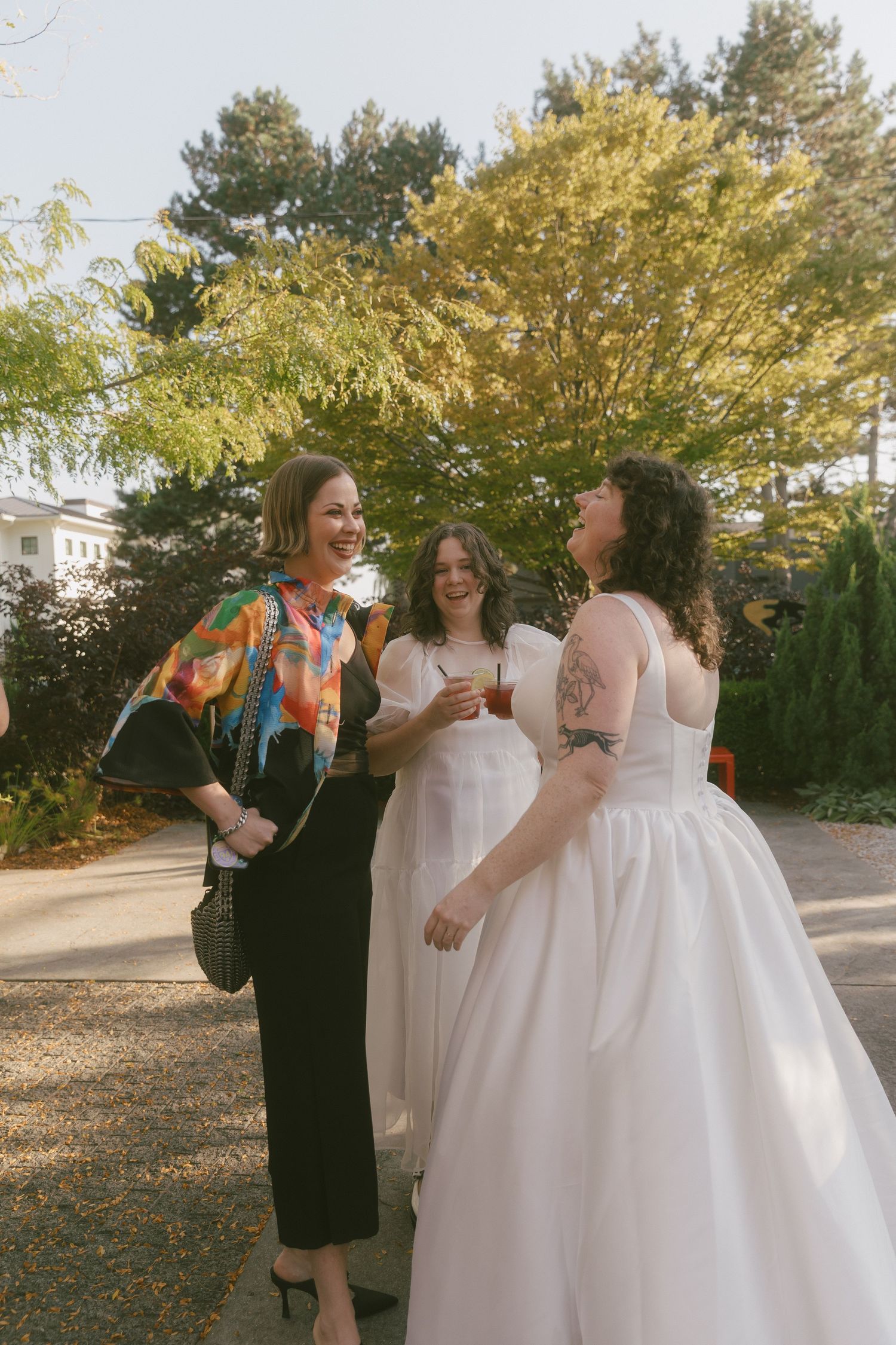 A group in formal attire laughs together at an outdoor wedding venue with trees in the background.