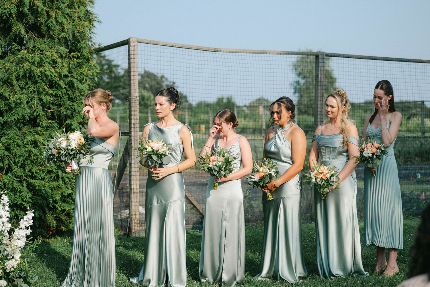 A group in sage green dresses stands together in a garden setting holding floral bouquets. - Elawa Farm