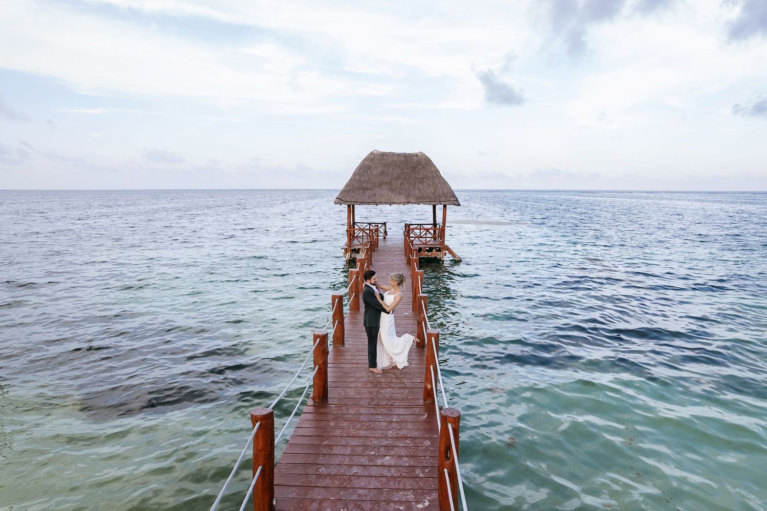 A long wooden pier stretches out into the turquoise ocean waters under a cloudy sky.