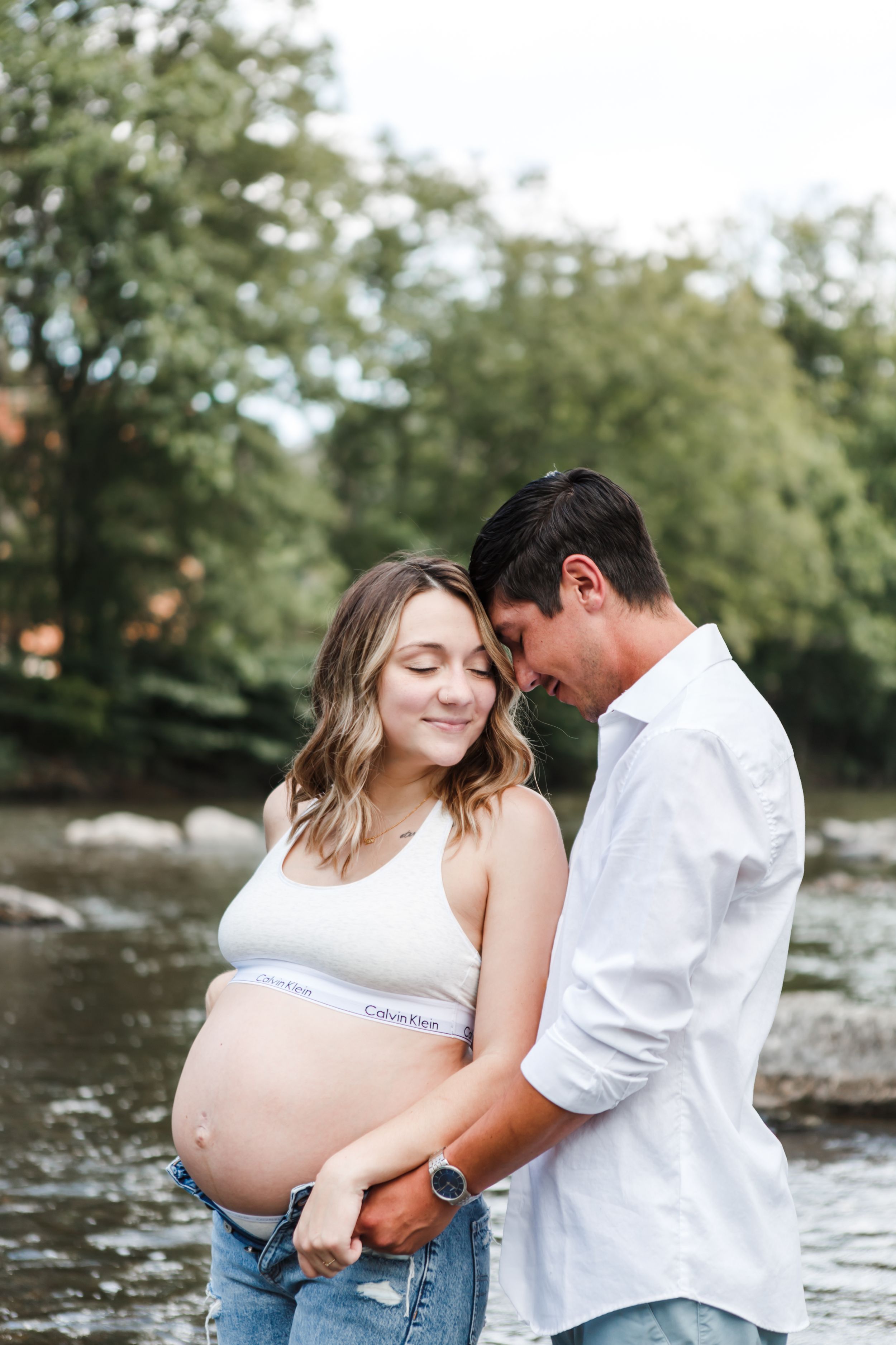 A couple shares an intimate moment by the water during a maternity photoshoot in a natural outdoor setting.