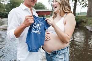 Couple in white shirts share coffee from blue mugs during outdoor maternity photoshoot by the water.