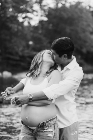 Black and white maternity photo of a romantic couple embracing outdoors by a lake.