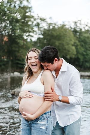 A couple shares intimate moments by the water during a maternity photoshoot in casual attire.