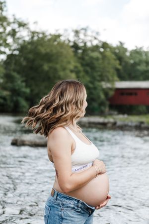 A maternity photo session by a river with a covered bridge visible in the background.