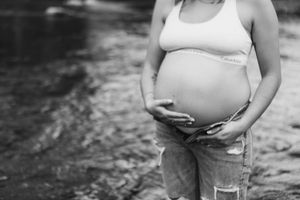 A couple stands together near water during an outdoor maternity photoshoot.