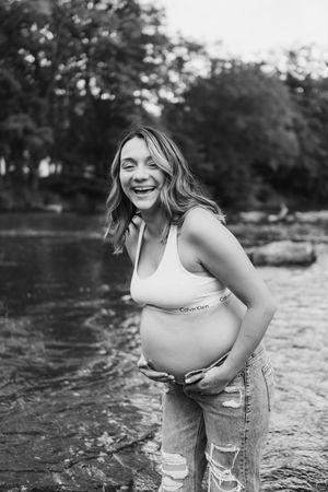 A pregnant woman in a white tank top stands in shallow water during a maternity photoshoot at a river.