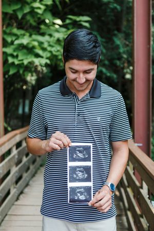 Person in striped shirt smiling while holding ultrasound photos on an outdoor boardwalk.