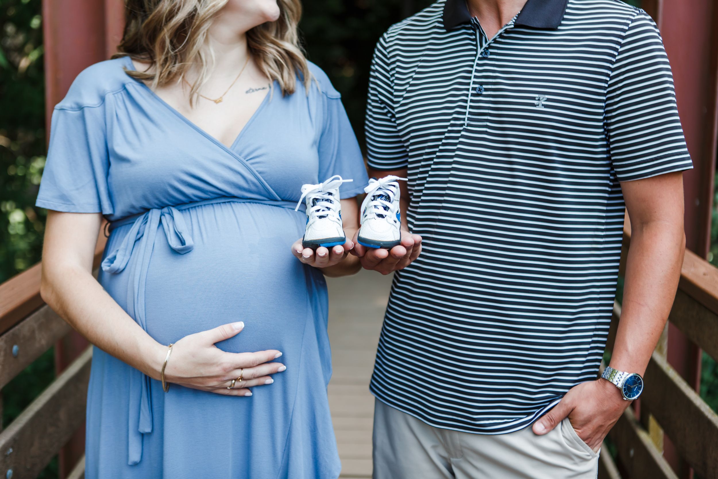 Maternity photos showing couple in coordinating blue outfits holding baby shoes by a wooden structure.
